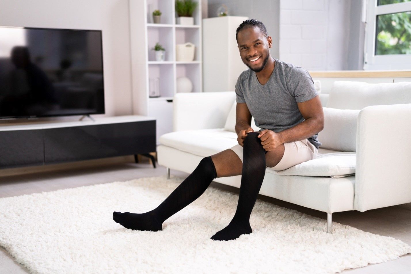 Man sitting on a white couch in a living room pulling up a pair of black compression socks