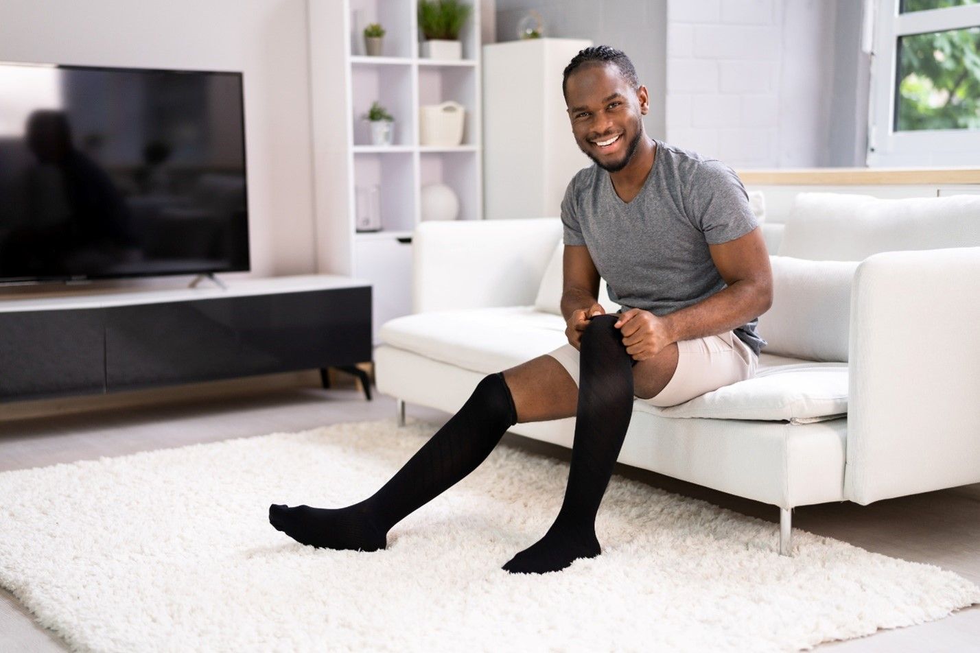 Man sitting on a white couch in a living room pulling up a pair of black compression socks 